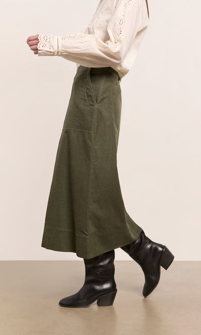 Plümo  Person wears a cream blouse, Beetle Green Skirt, and black heeled boots while walking indoors.