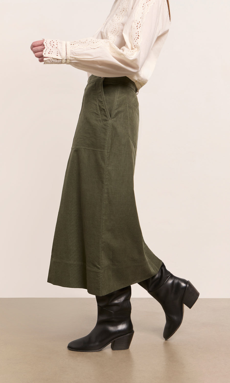 Plümo  Person wears a cream blouse, Beetle Green Skirt, and black heeled boots while walking indoors.