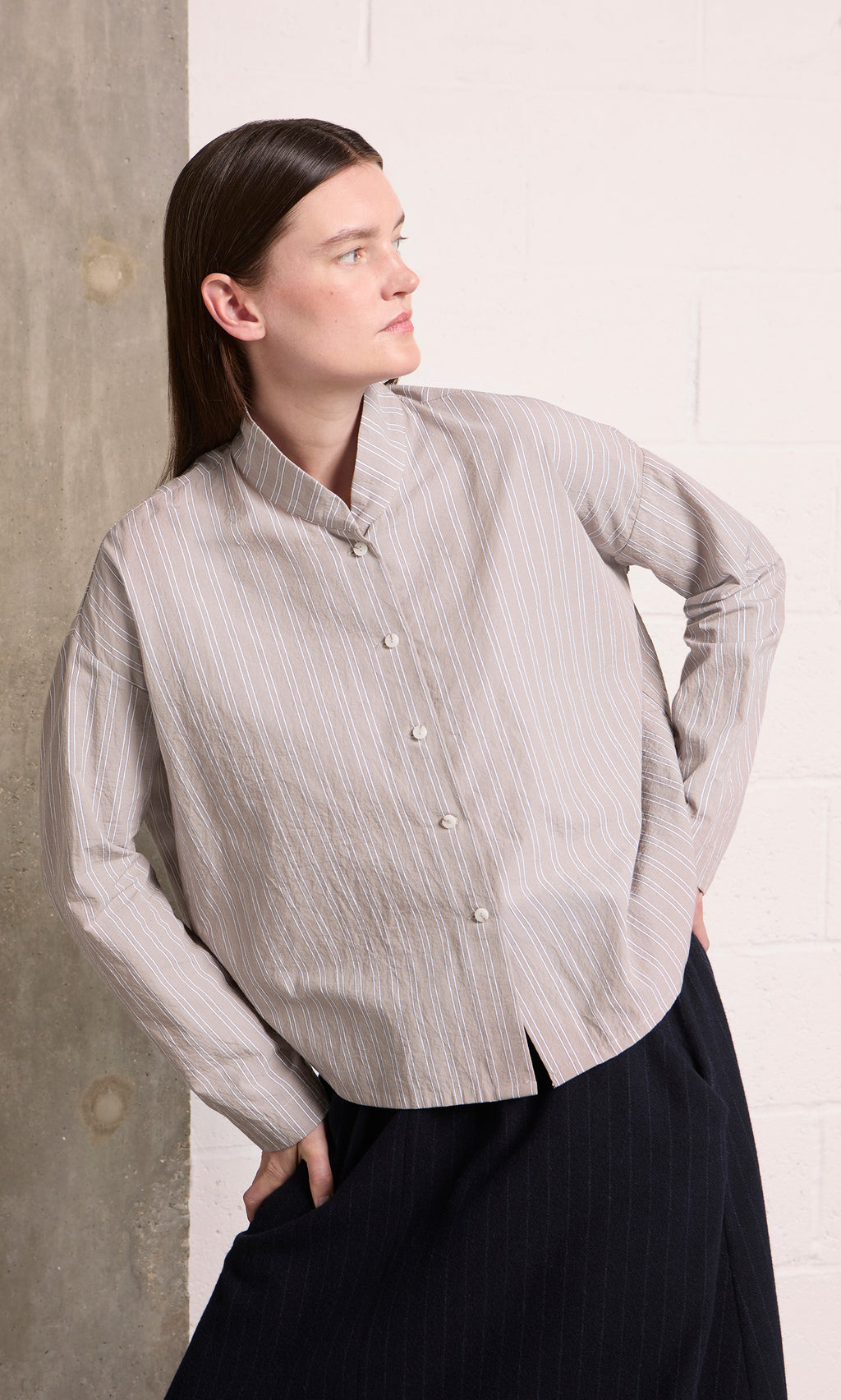 Plümo  Person in the Bennett Blouse - Smoke stands by a concrete wall, looking left with one hand on their hip.