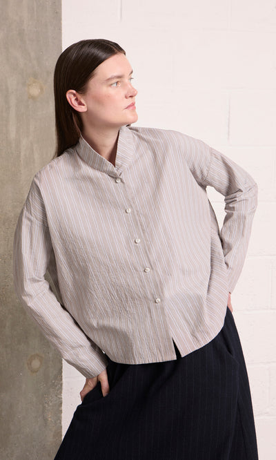 Plümo  Person in the Bennett Blouse - Smoke stands by a concrete wall, looking left with one hand on their hip.