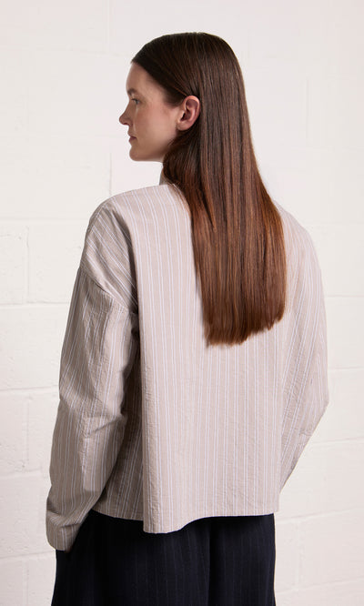 Plümo  Person with straight brown hair wears the Bennett Blouse - Smoke, facing away against a white brick wall.