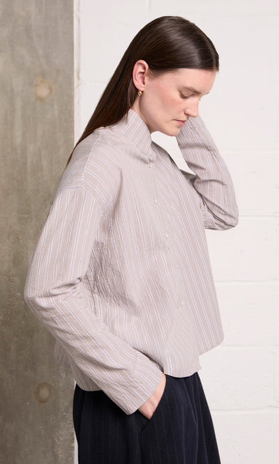 Plümo  A woman in the Bennett Blouse - Smoke faces sideways, touching her hair against a textured wall.