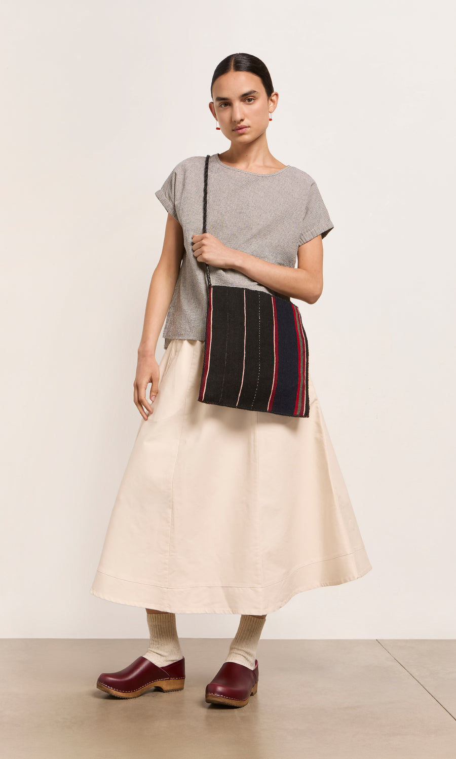Plümo  A woman wears a grey top, cream skirt, Low Jord Clogs, and carries a striped bag against a white background.