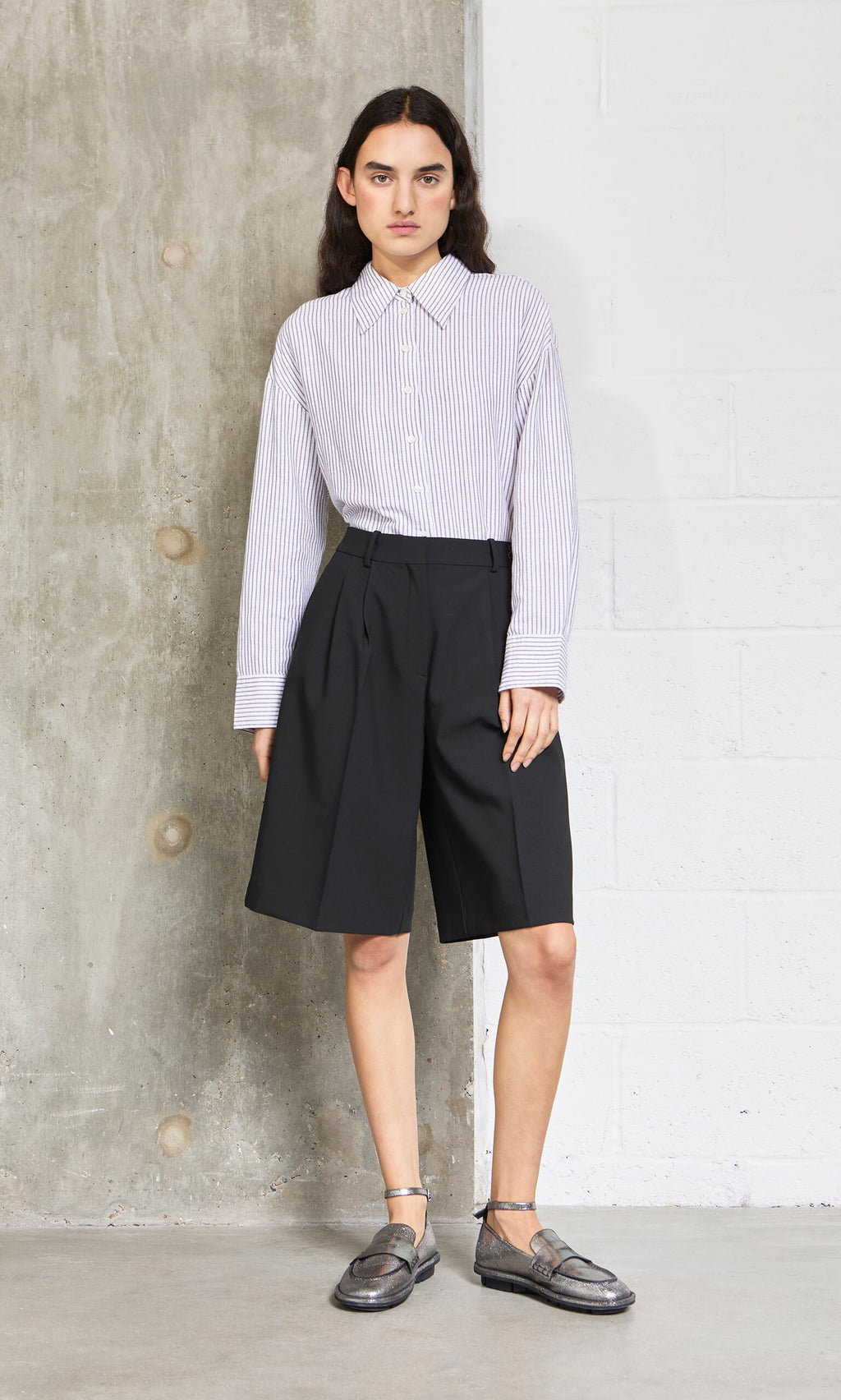 Plümo- Person stands against a concrete wall wearing an oversized Boswell Shirt, black knee-length shorts, and loafers.