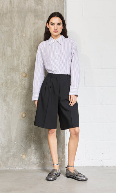 Plümo- Person stands against a concrete wall wearing an oversized Boswell Shirt, black knee-length shorts, and loafers.