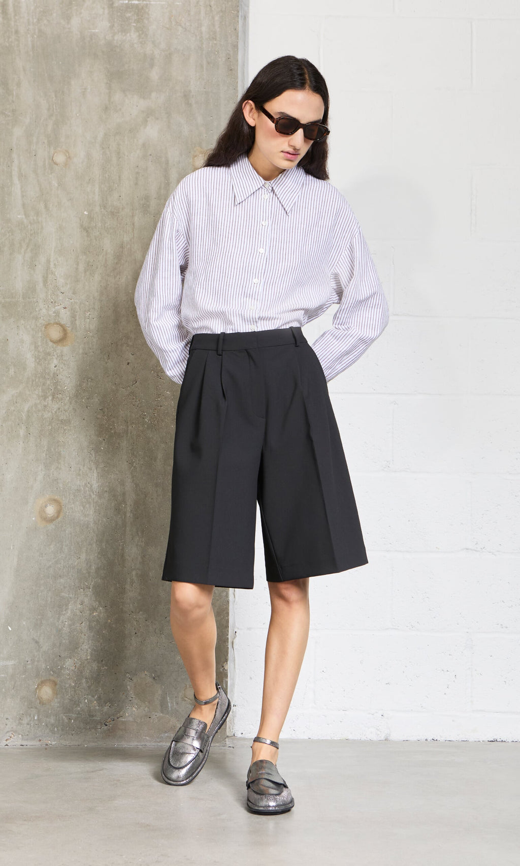 Plümo- Person wears Bermuda Shorts, a striped button-up shirt, metallic shoes, and sunglasses standing against a wall.
