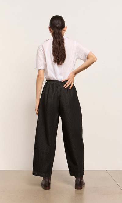 Plümo  Person with long dark hair in a ponytail wears a white shirt and Black Barrel Pants, standing with back facing.