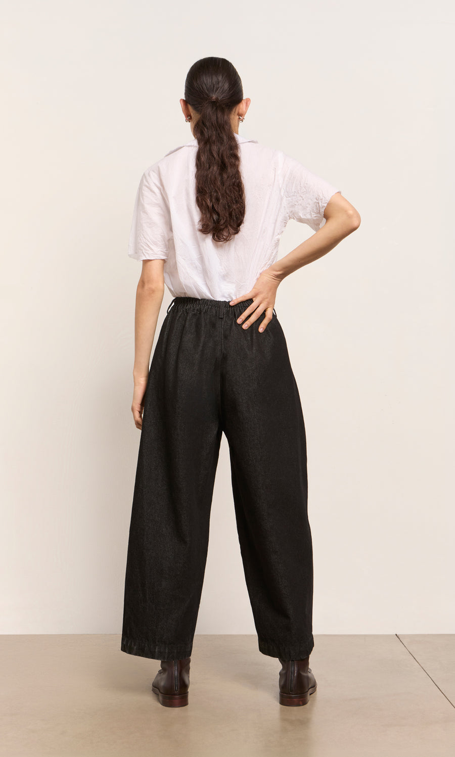 Plümo  Person with long dark hair in a ponytail wears a white shirt and Black Barrel Pants, standing with back facing.