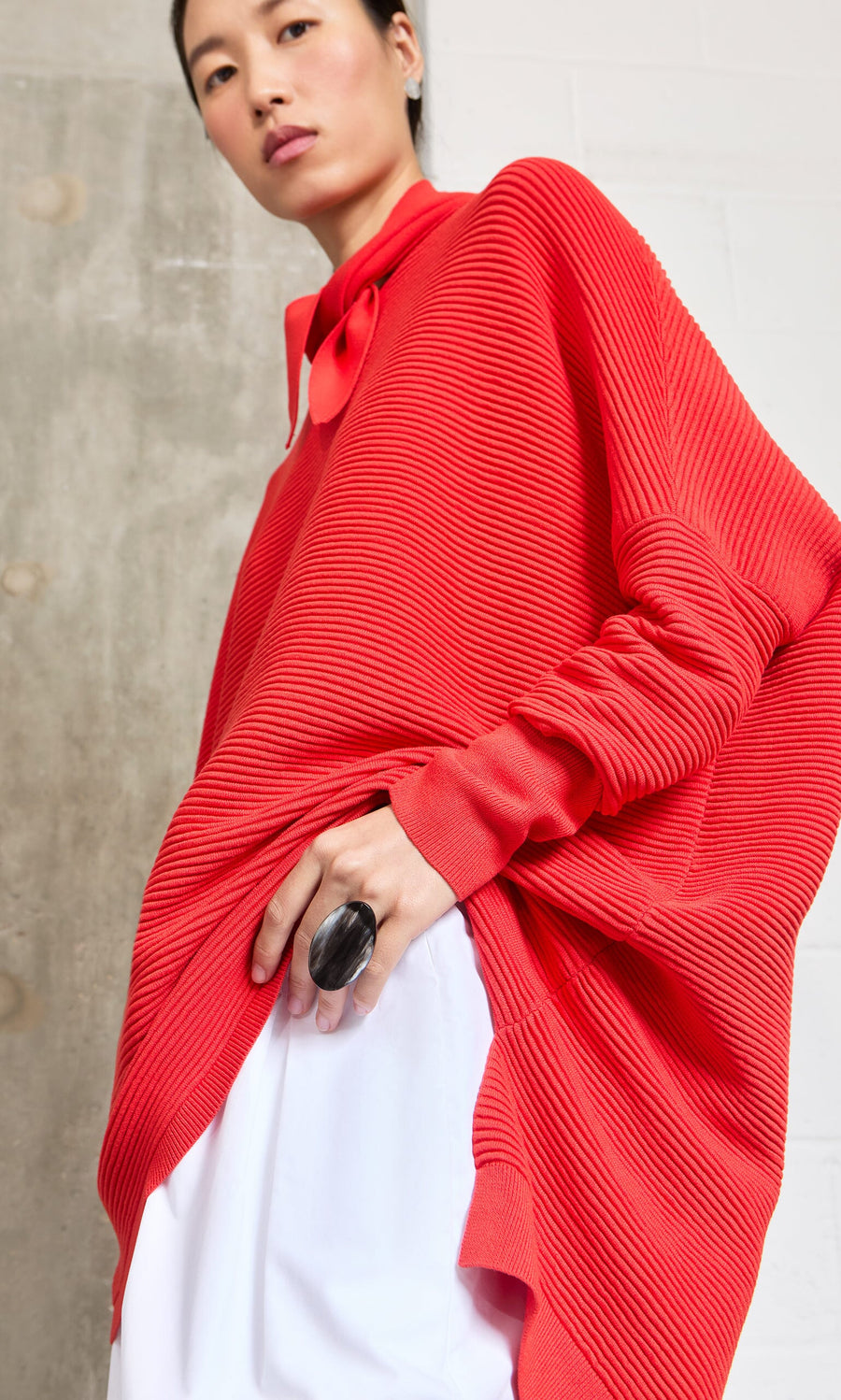 Plümo- Person wearing an oversized red textured top, white pants, and a Black Oval Ring, standing against a concrete wall.