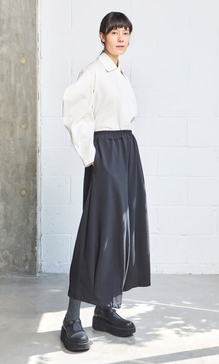 Plümo- Person standing indoors wearing a white Ting Blouse with bishop sleeves, dark wide-leg pants, socks, and black shoes.
