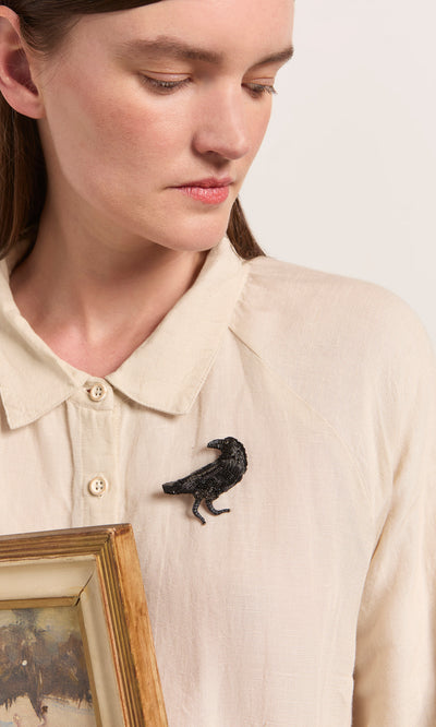 Plümo  Woman wearing a cream shirt and the Black Raven Brooch, holding a framed picture and looking down.