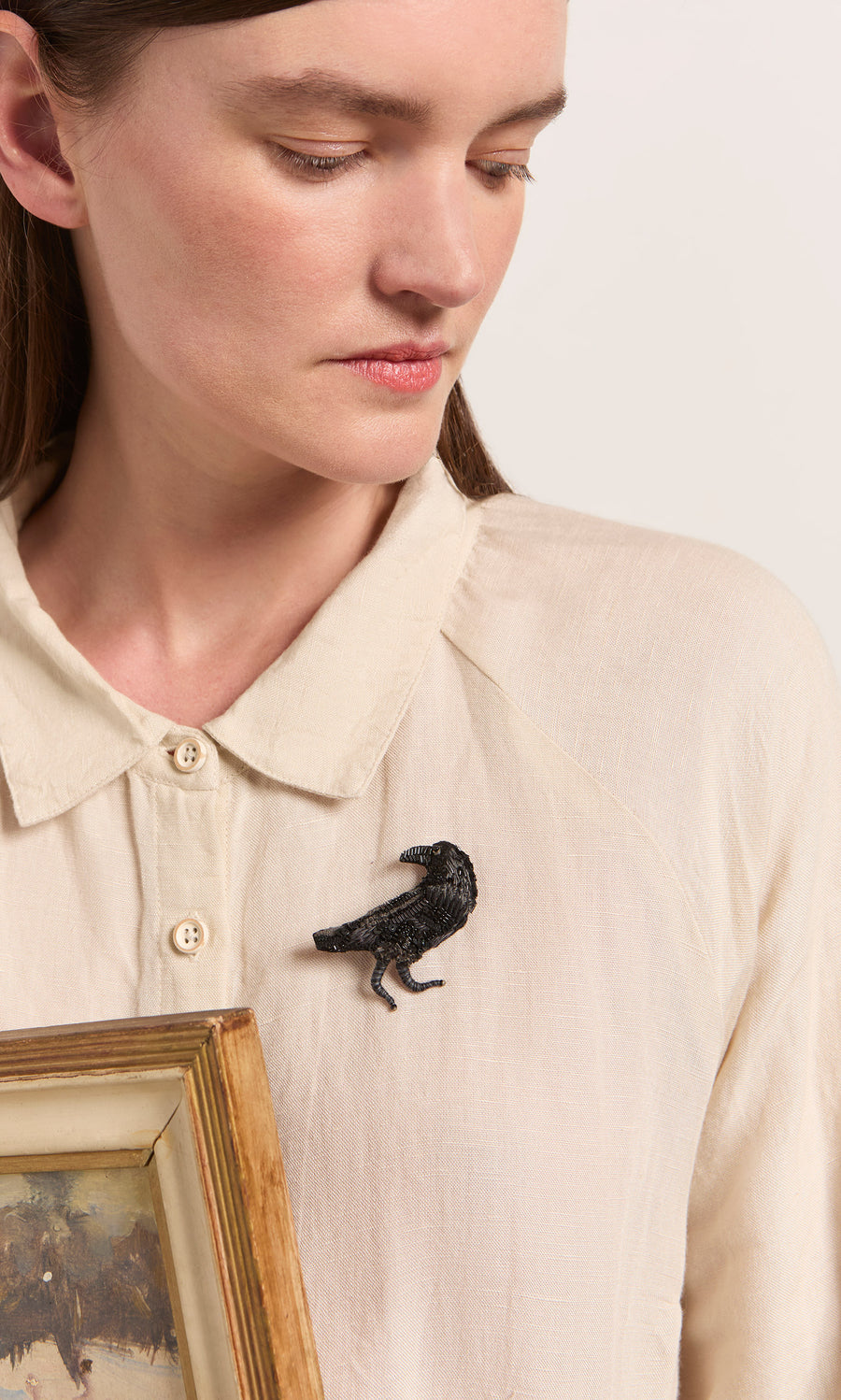 Plümo  Woman wearing a cream shirt and the Black Raven Brooch, holding a framed picture and looking down.