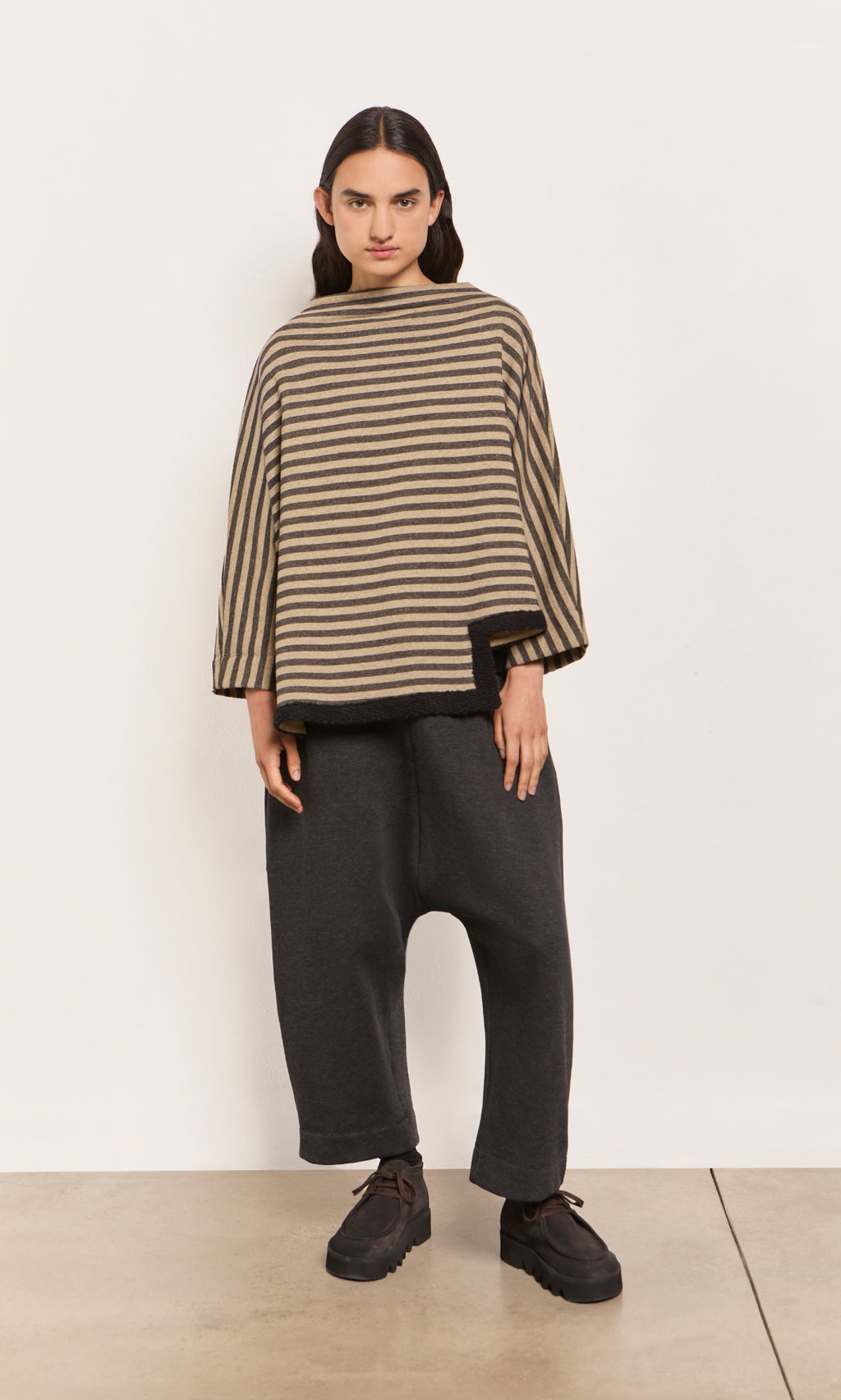 Plümo  Person wears the Bloch Funnel Top, featuring taupe and black stripes with an asymmetric cut-out hem, plus loose dark pants.