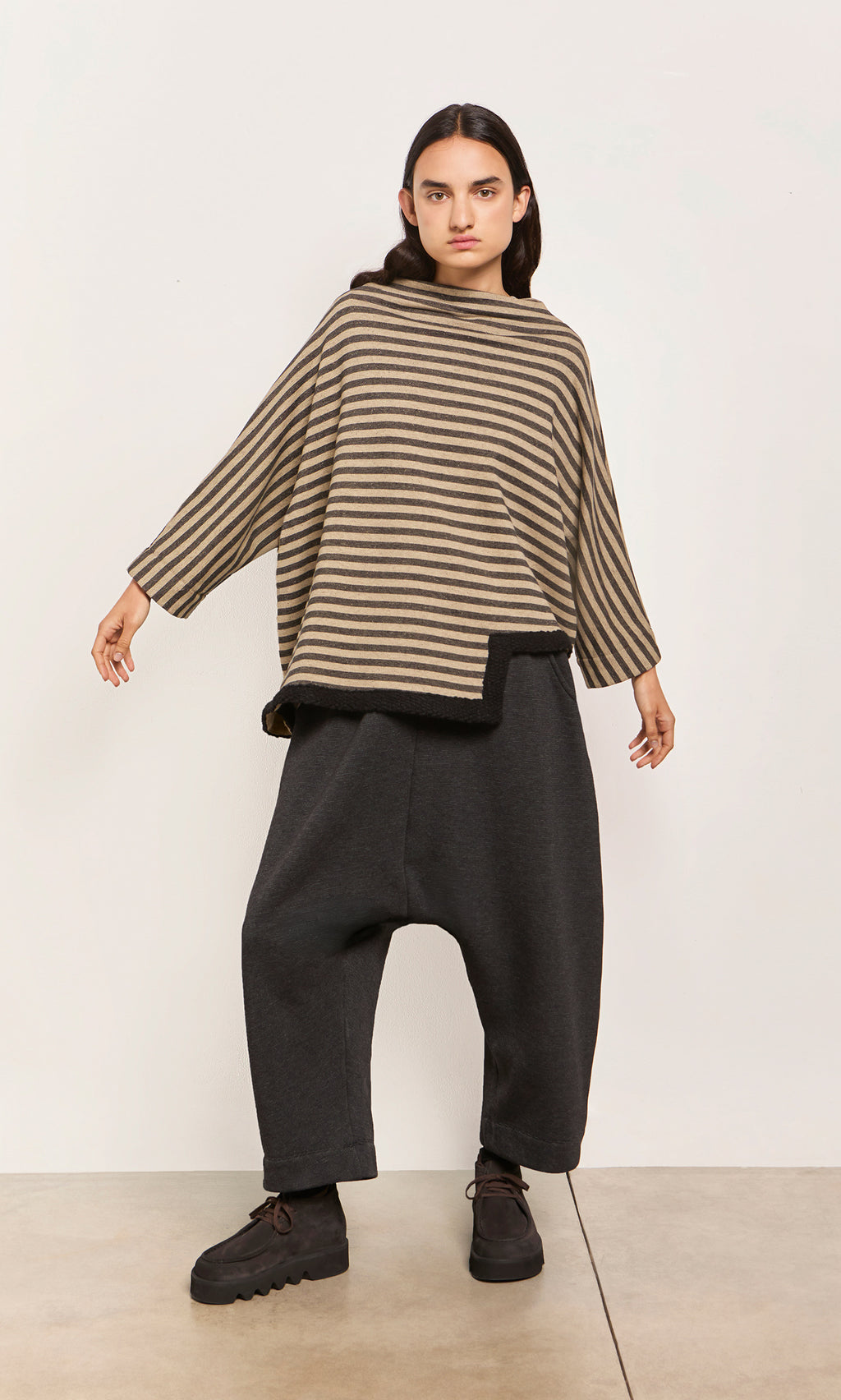 Plümo  Person wears the Bloch Funnel Top in taupe/black stripes with boucle trim, baggy dark pants, arms outstretched by a wall.