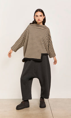 Plümo  Person wears the Bloch Funnel Top in taupe/black stripes with boucle trim, baggy dark pants, arms outstretched by a wall.