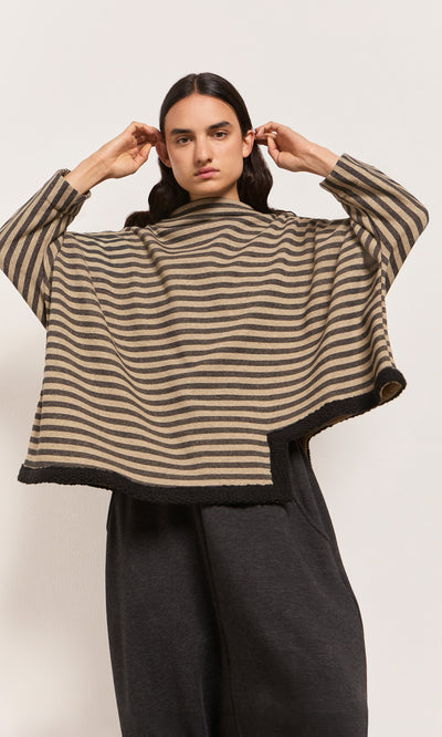 Plümo  Person in the Bloch Funnel Top, taupe/black stripes with wide sleeves, boucle trim, and dark pants, adjusts hair.