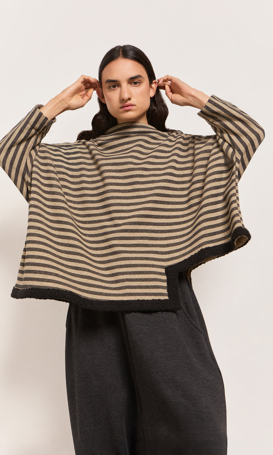 Plümo  Person in the Bloch Funnel Top, taupe/black stripes with wide sleeves, boucle trim, and dark pants, adjusts hair.