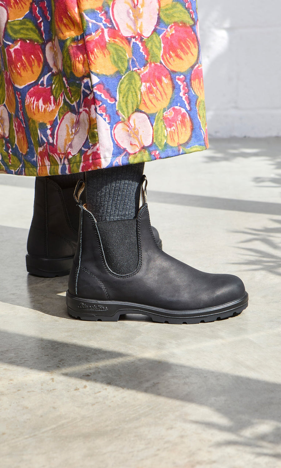 Plümo- Person wearing Blundstone Boots - Black, dark socks, and a colorful floral skirt on a light concrete floor.