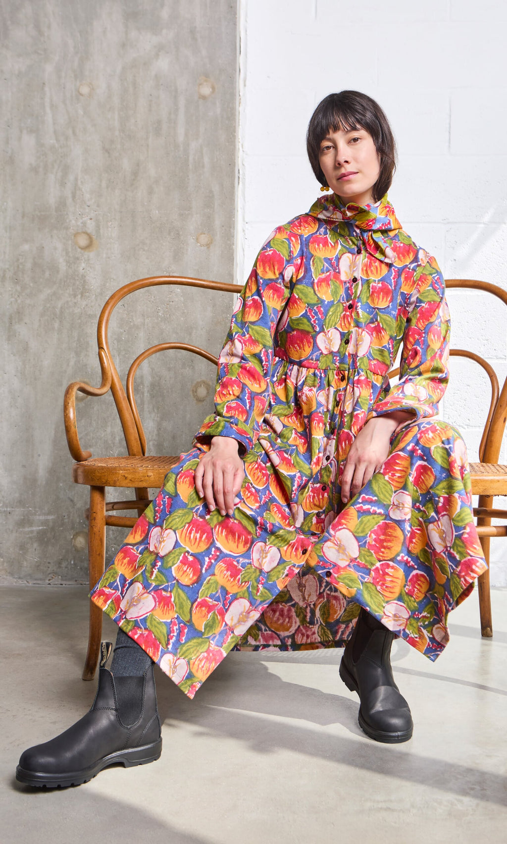 Plümo- Person in a Braeburn Dress with rosy apple print and black boots sits on a wooden bench in a minimal, modern setting.