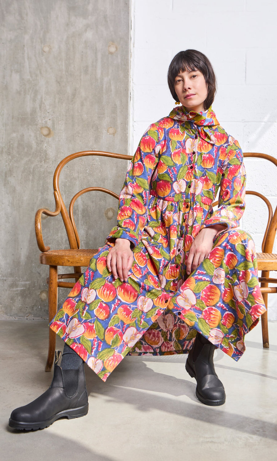 Plümo- Person in a Braeburn Dress with rosy apple print and black boots sits on a wooden bench in a minimal, modern setting.