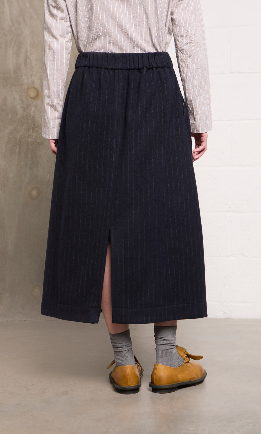 Plümo  Person wears Bo Skirt—navy wool pinstripe, midi with back slit—plus gray socks and yellow shoes, standing indoors.