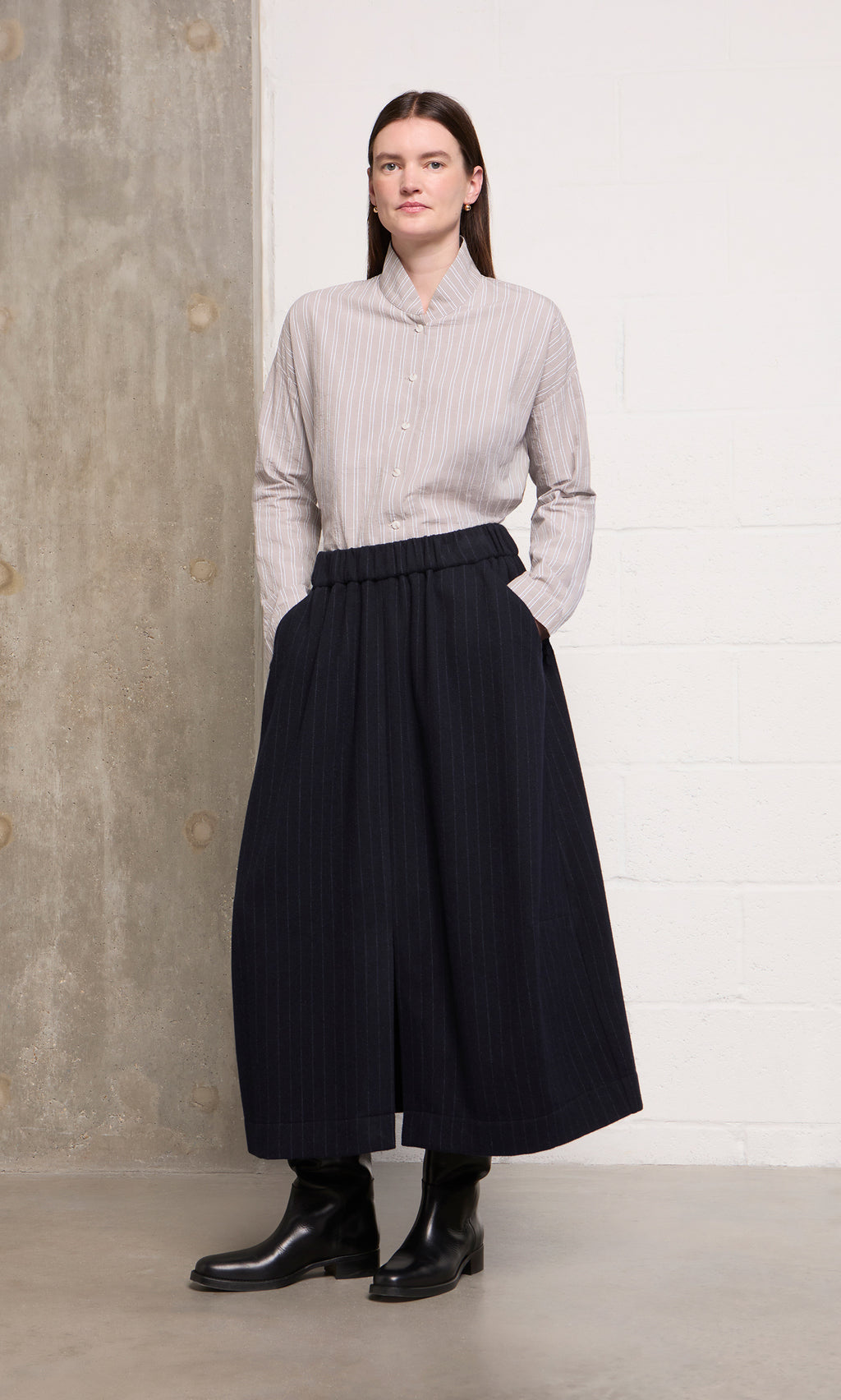 Plümo  Woman in a striped shirt, Bo Skirt, and black boots stands by a concrete and white wall.