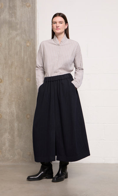 Plümo  Woman in a striped shirt, Bo Skirt, and black boots stands by a concrete and white wall.