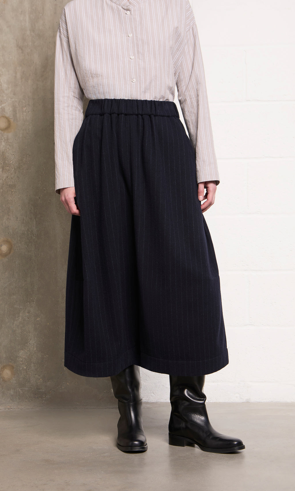 Plümo  Model in beige striped top, Bo Skirt in wool pinstripe, and black boots stands against a wall.