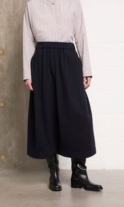 Plümo  Model in beige striped top, Bo Skirt in wool pinstripe, and black boots stands against a wall.