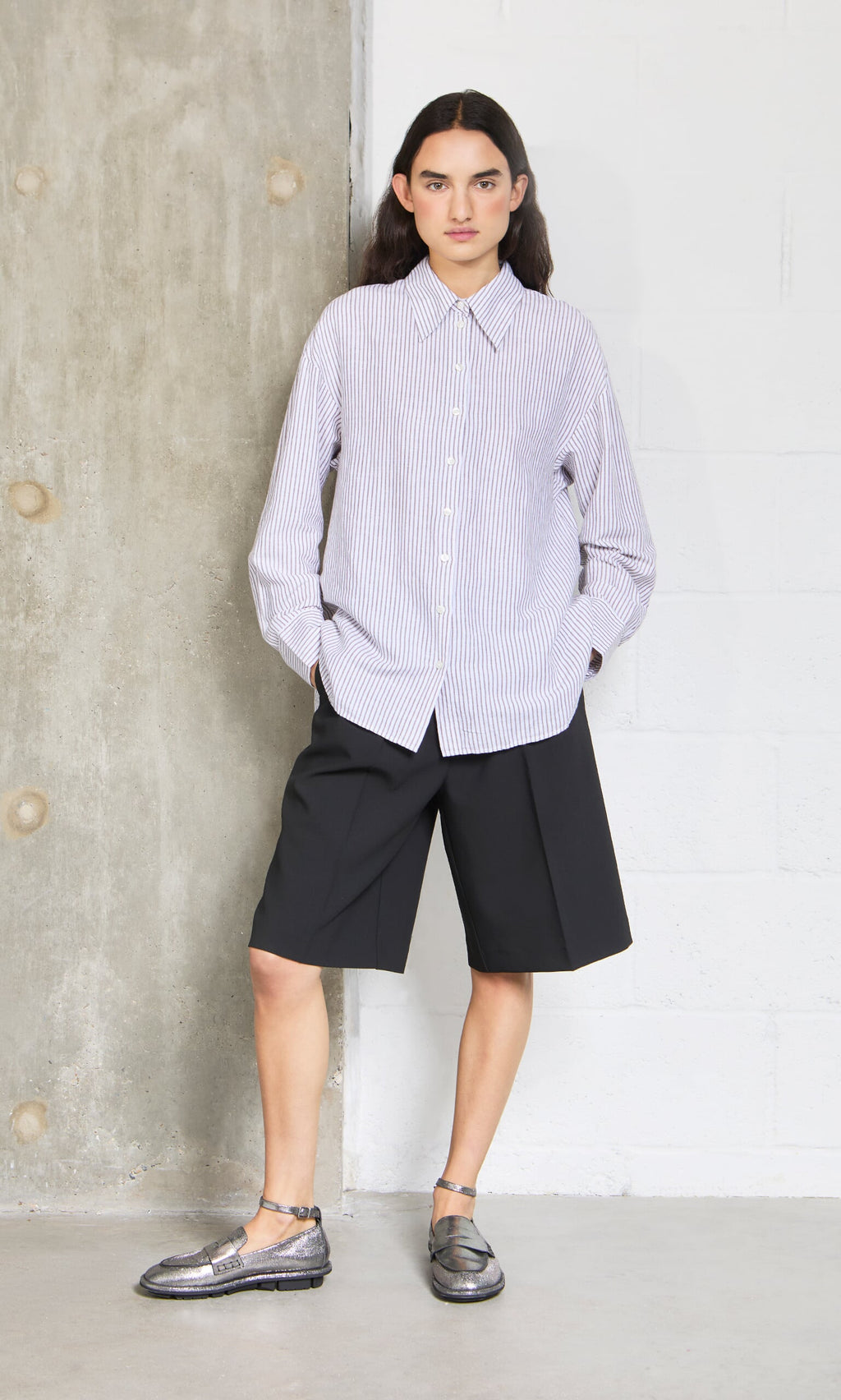Plümo- Person wears the Boswell Shirt with black knee-length shorts and metallic shoes, standing by a textured wall.