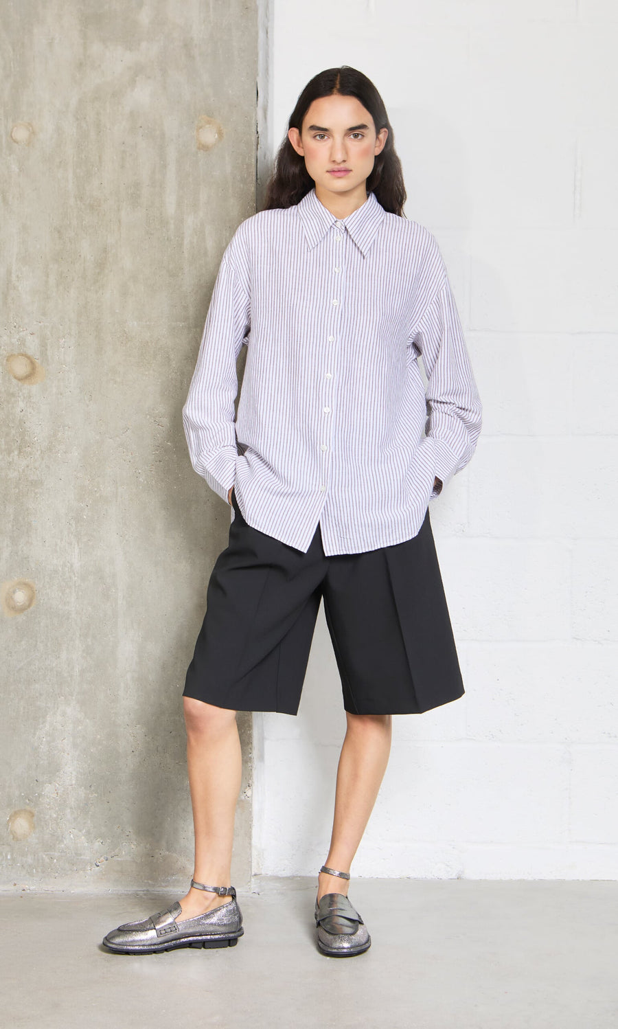 Plümo- Person wears the Boswell Shirt with black knee-length shorts and metallic shoes, standing by a textured wall.