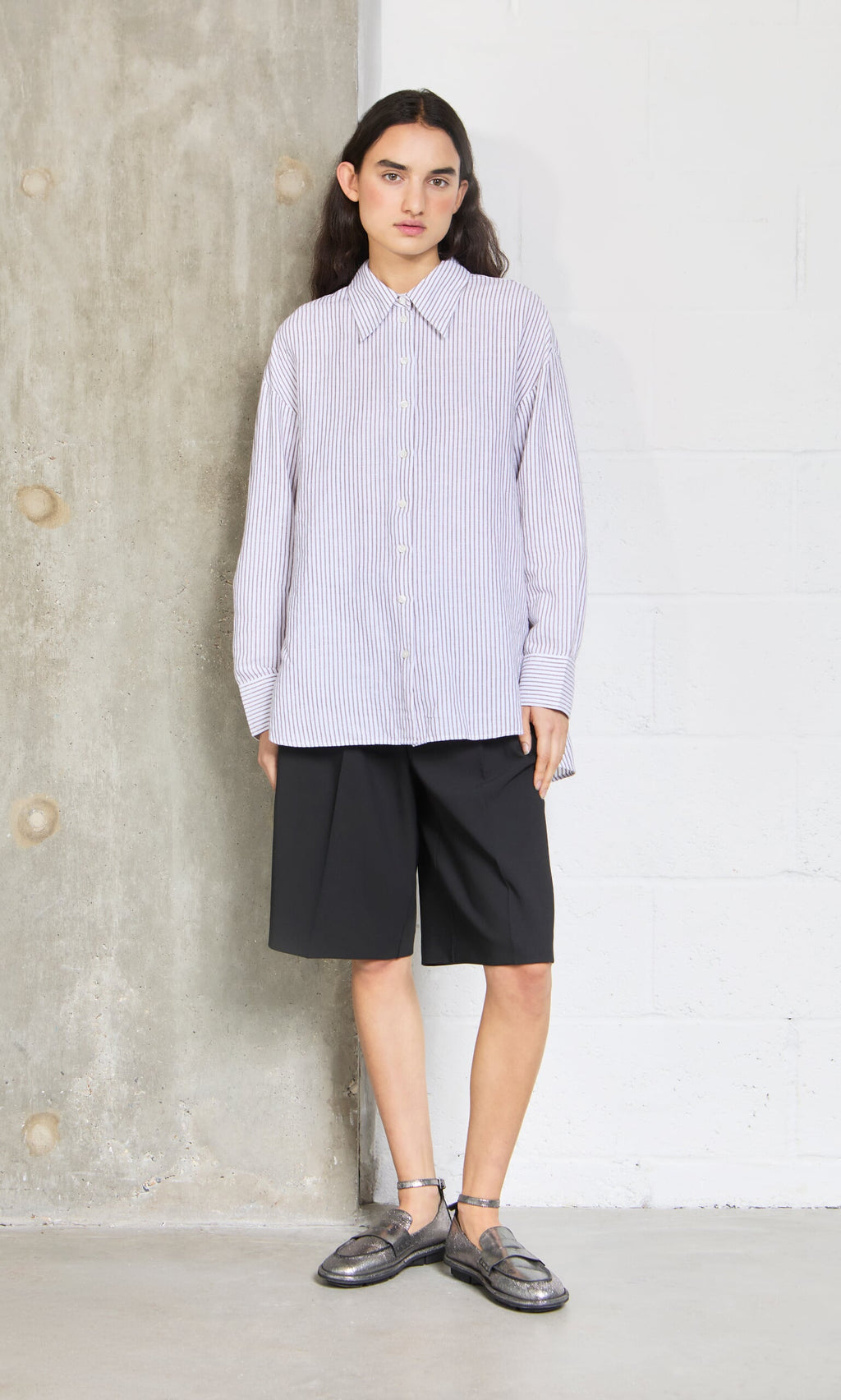 Plümo- Person wearing the Boswell Shirt, black knee-length shorts, and loafers, standing against a concrete and white wall.