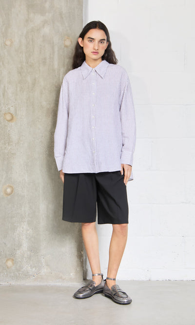 Plümo- Person wearing the Boswell Shirt, black knee-length shorts, and loafers, standing against a concrete and white wall.