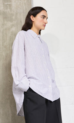 Plümo- A person wears the oversized Boswell Shirt and dark pants, standing sideways with hands in pockets.