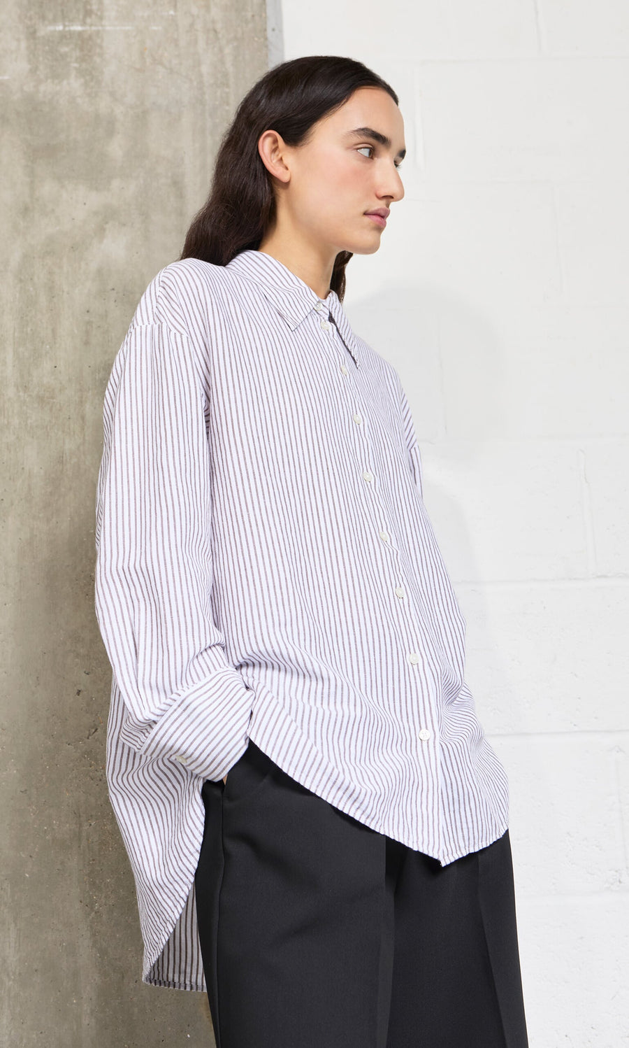 Plümo- A person wears the oversized Boswell Shirt and dark pants, standing sideways with hands in pockets.