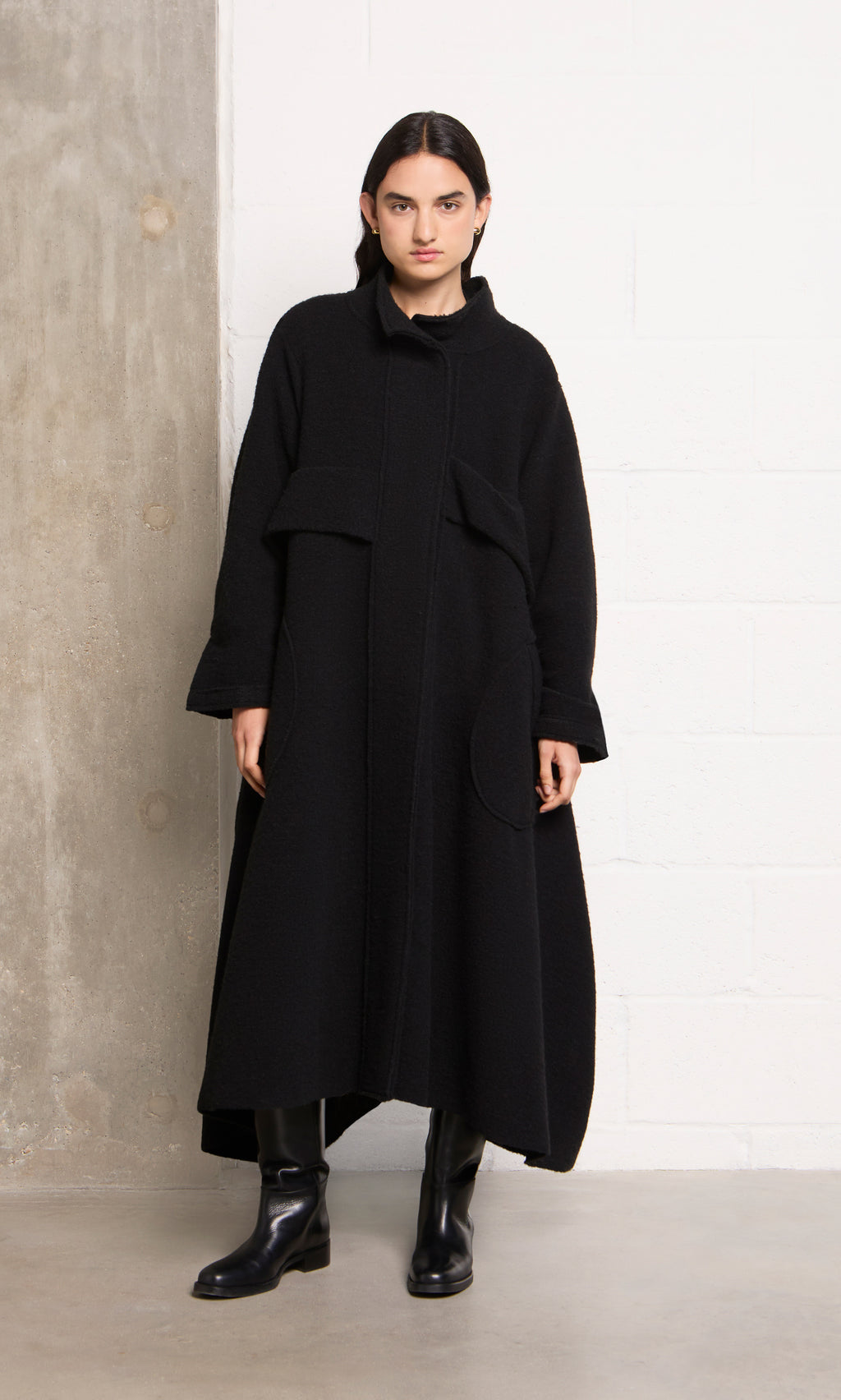 Plümo  Person in the black Brabant Coat and boots stands against a concrete and white brick wall.