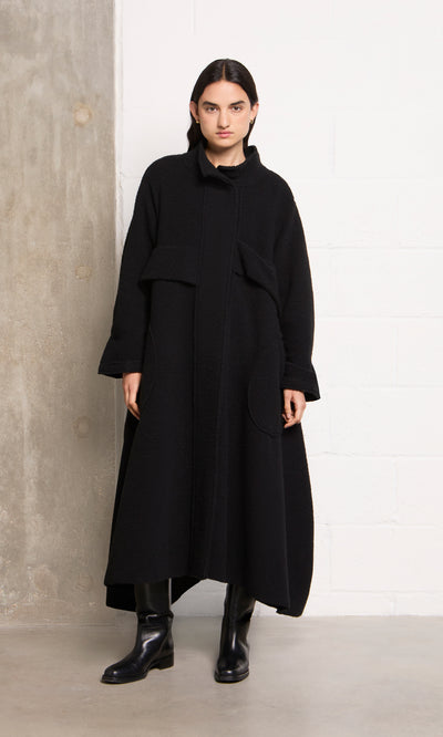 Plümo  Person in the black Brabant Coat and boots stands against a concrete and white brick wall.
