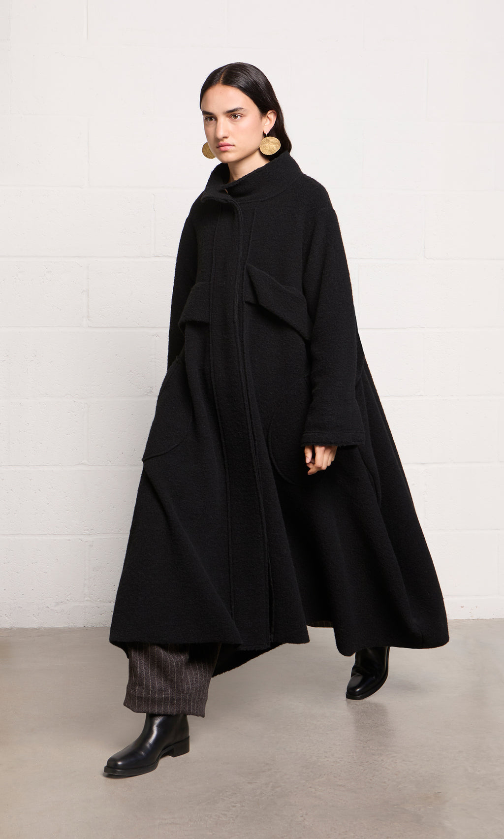 Plümo  Woman in a Brabant Coat—long, black boucle wool with large pockets—plus dark pants and black boots, walking indoors.