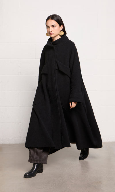 Plümo  Woman in a Brabant Coat—long, black boucle wool with large pockets—plus dark pants and black boots, walking indoors.