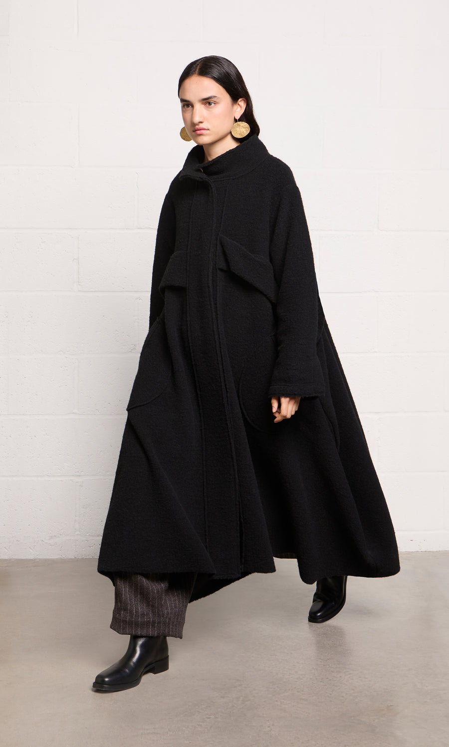 Plümo  Woman in a Brabant Coat—long, black boucle wool with large pockets—plus dark pants and black boots, walking indoors.