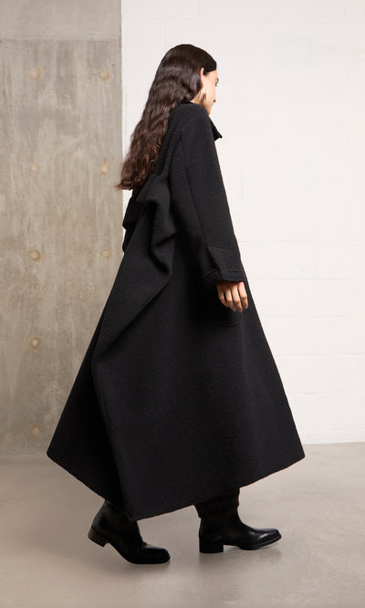 Plümo  Person in the Brabant Coat and boots walks indoors by a concrete and white wall, facing left.
