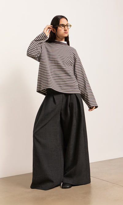 Plümo  Woman in a striped top and Pleated Grey Pants stands on a light floor, wearing glasses, looking to the side.