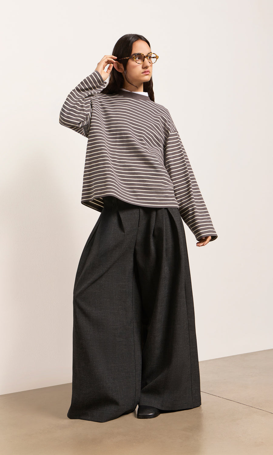 Plümo  Woman in the Breton Tee - Coal and black wide-leg pants stands by a plain wall, adjusting her glasses.