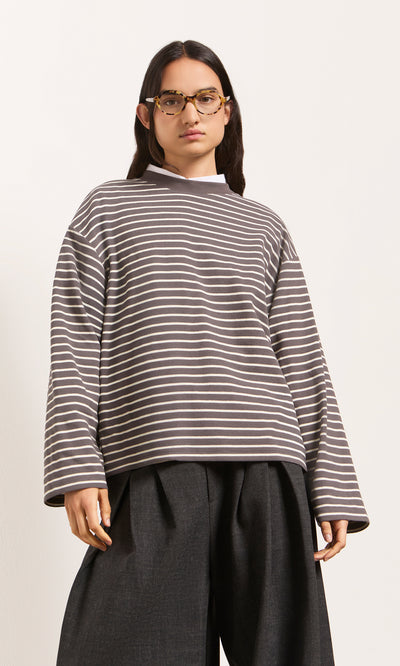 Plümo  Wearing the Breton Tee - Coal, wide dark pants, and glasses, someone poses against a plain background.