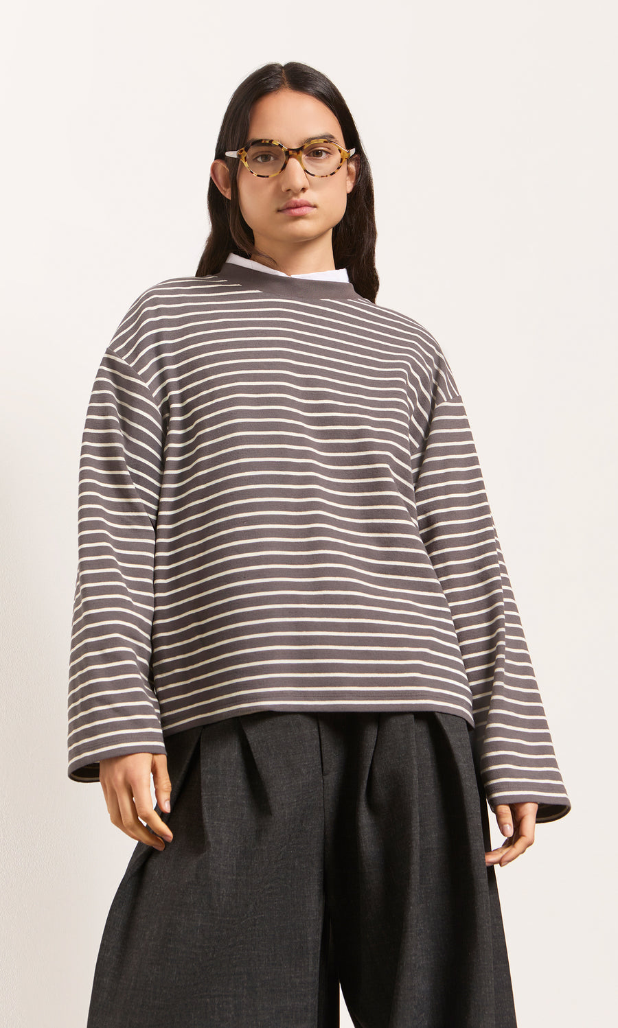 Plümo  Wearing the Breton Tee - Coal, wide dark pants, and glasses, someone poses against a plain background.