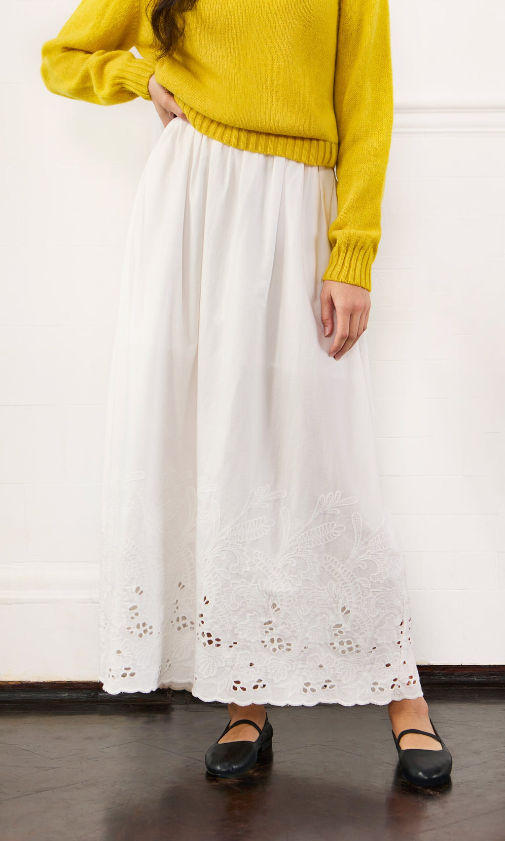 Plümo- Person in yellow sweater and Bright Skirt stands against white wall, wearing black Mary Jane shoes.