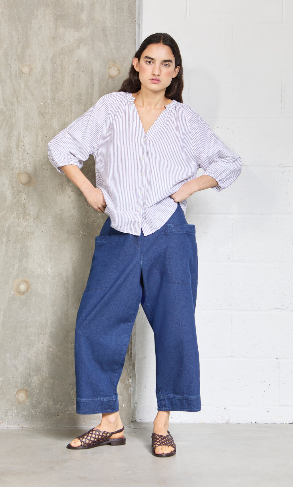 Plümo- Woman stands with hands in relaxed fit blue jeans pockets, wearing the Brisley Blouse and sandals by a concrete wall.