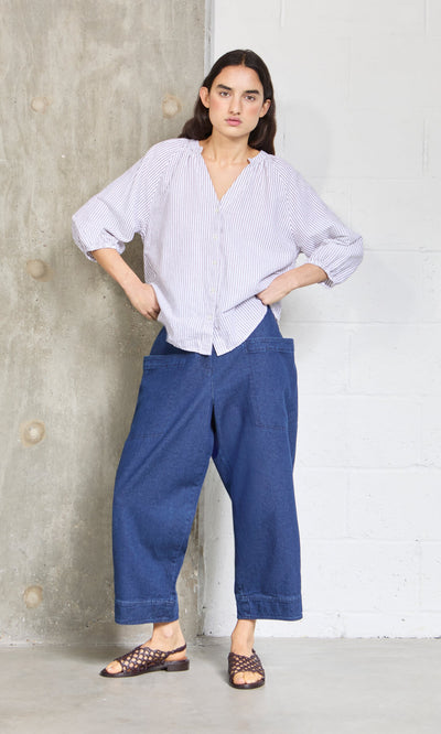 Plümo- Woman stands with hands in relaxed fit blue jeans pockets, wearing the Brisley Blouse and sandals by a concrete wall.