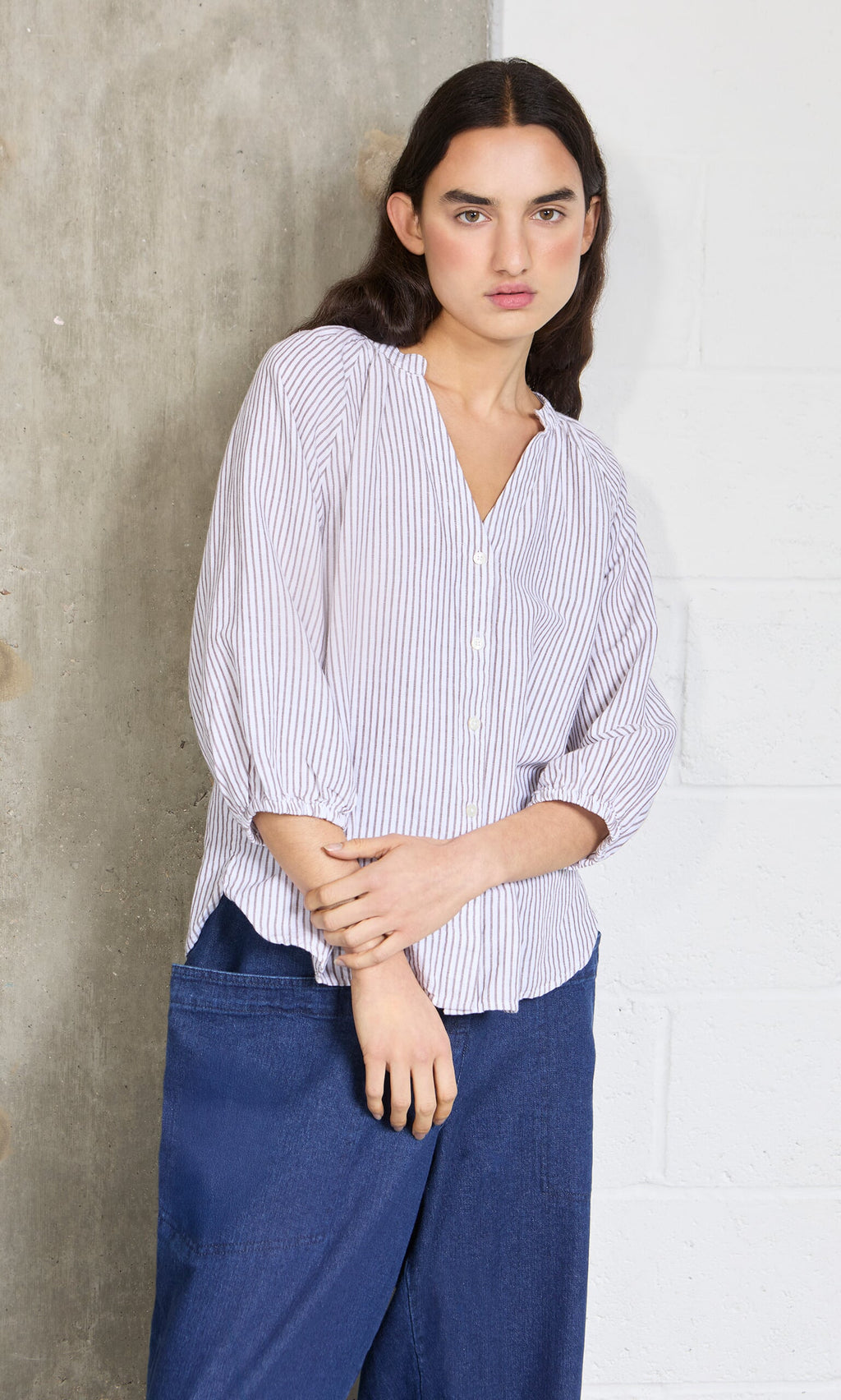 Plümo- Wearing the Brisley Blouse and blue pants, a person stands by a concrete and white brick wall, looking at the camera.