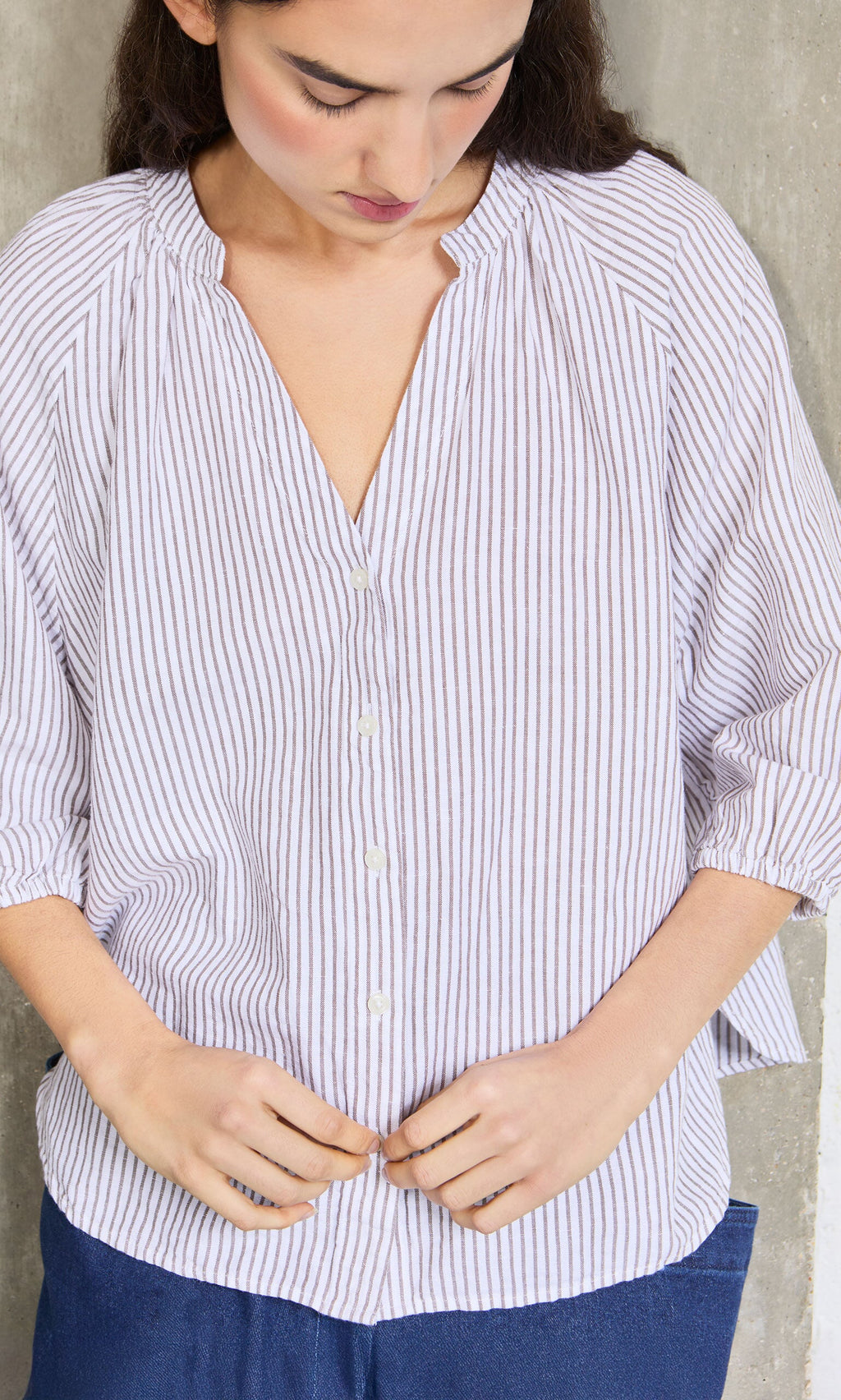 Plümo- Woman in a relaxed fit Brisley Blouse, looking down as she buttons her shirt against a concrete wall.